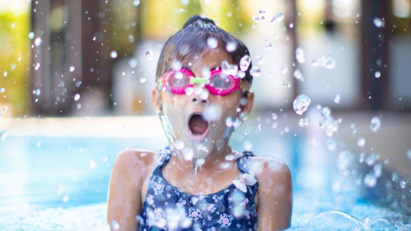 Menina com cerca de 6 anos, usando óculos de natação infantil rosa, mergulhando numa piscina e espelhando água.