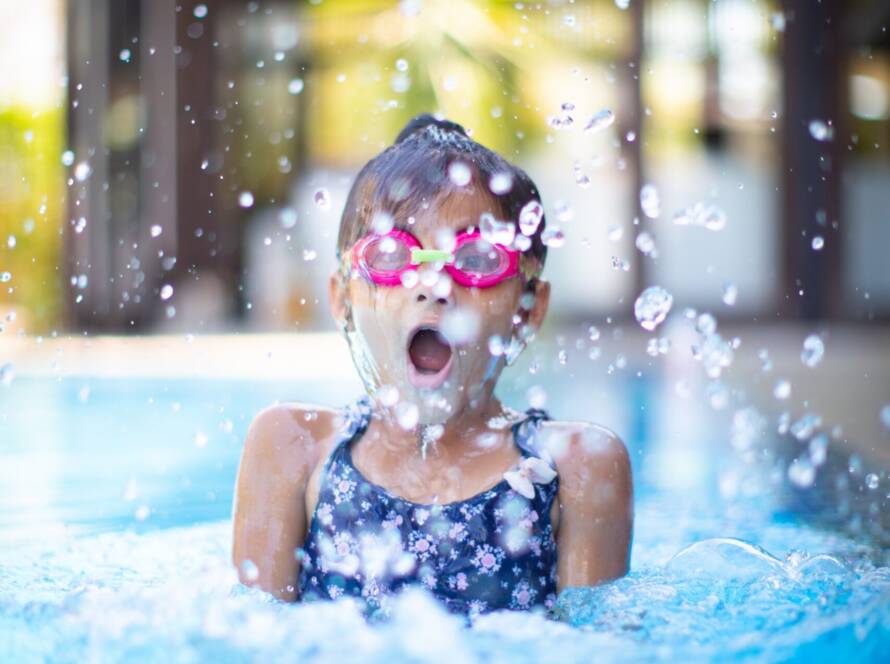 Menina com cerca de 6 anos, usando óculos de natação infantil rosa, mergulhando numa piscina e espelhando água.